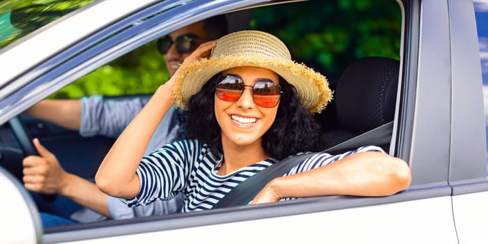 Ecstatic millennial arab husband and wife having car trip, waving from window, shot from outside, side view. Happy young lady going vacation with her boyfriend, wearing sunglasses and summer hat