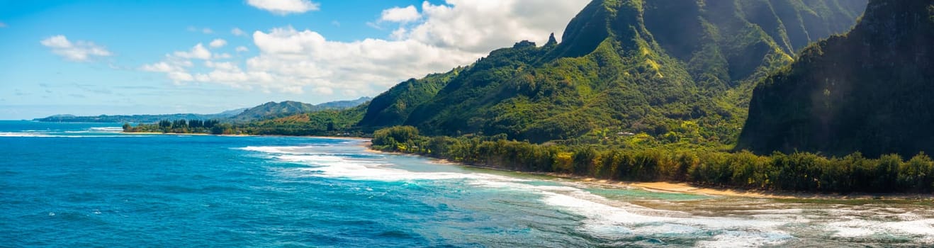 Aerial view of Kauai Island, Hawaii, showing turquoise waters, sandy beach, lush Napali cliffs, and scattered clouds under a vibrant blue sky.