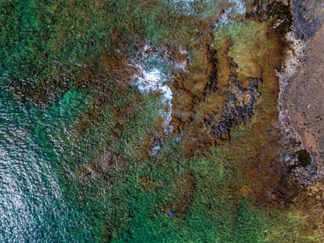 Aerial perspective of Kauai Island's coastline with turquoise waters, volcanic rocks, golden brown sand, and white waves breaking along the shore.