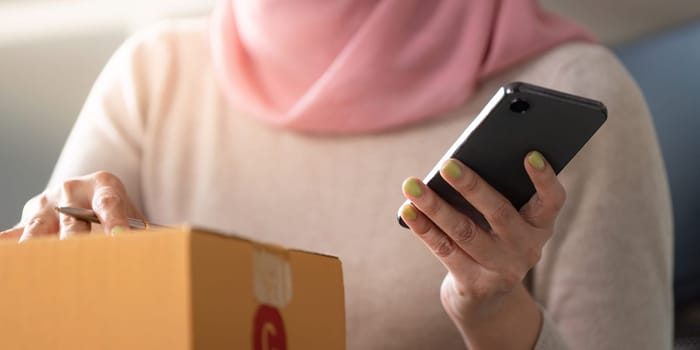 A close-up view of a Muslim woman in a hijab checking her smartphone while preparing a package for an online order, showcasing modern entrepreneurship.