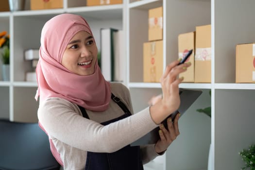 A Muslim woman in a hijab and apron smiling while managing her inventory for online sales using a tablet in her organized home office, reflecting her entrepreneurial spirit.
