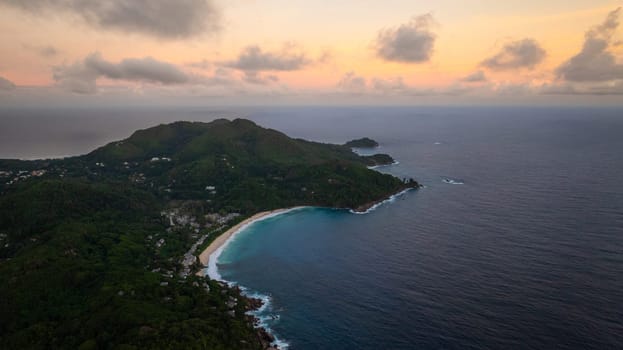 Sunset view of a tropical coastline with lush green hills, calm ocean waters, and a soft pink sky. Seychelles, Mahe.