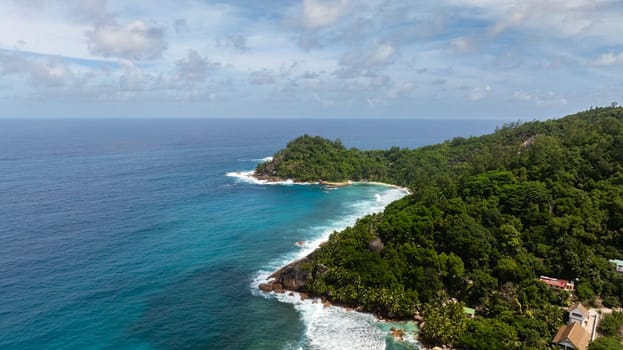 Cliffside ocean view with turquoise waters meeting lush greenery, with waves crashing on the rocky shore. Seychelles, Mahe.