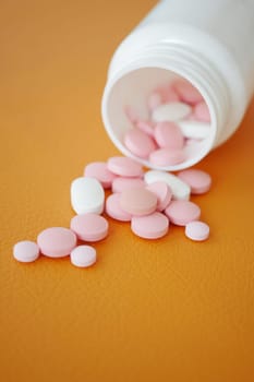 Various pink and white pills scattered from a white bottle on orange.