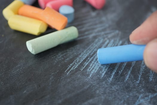 A hand draws with blue chalk on a dark surface, surrounded by colorful chalks.