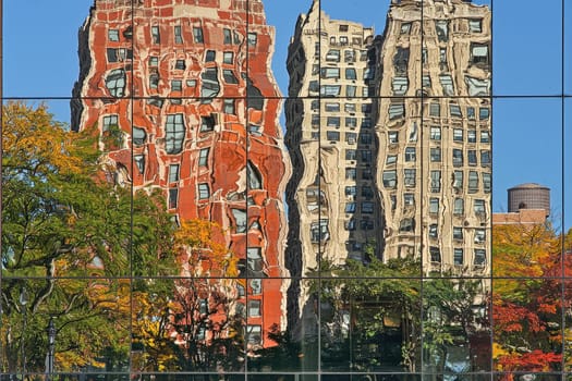 Two tall buildings, one of which has a red facade. The other building has a brown facade. The red building is reflected in the window of the brown building, creating a beautiful and unique scene