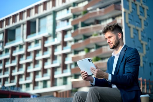 Young businessman analyzing work on his digital tablet while sitting outside a modern office building.