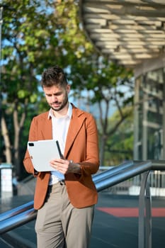 Handsome businessman using digital tablet, leaning on a railing outside contemporary glass office.
