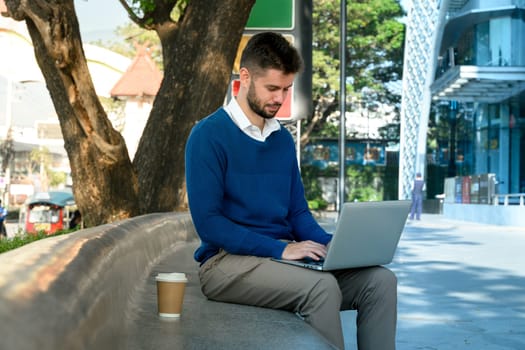 Caucasian male freelancer working on his laptop sitting on a curved outdoor bench.