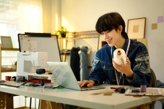 Smiling designer holding a spool of thread working on laptop in a stylish workshop.