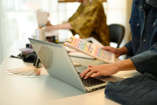 Fashion designer selecting fabric colors while working on laptop in a modern studio.