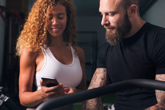 latin woman with curly hair in white top showing smartphone to man with tattoos in room, concept of technology of communication
