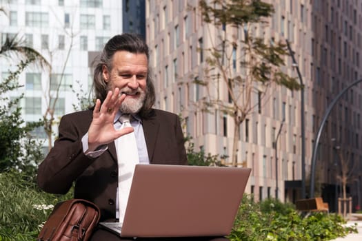 happy successful mature male in elegant suit waving hand and saying hello while having online video call via laptop sitting on urban square, concept of business and middle age man lifestyle