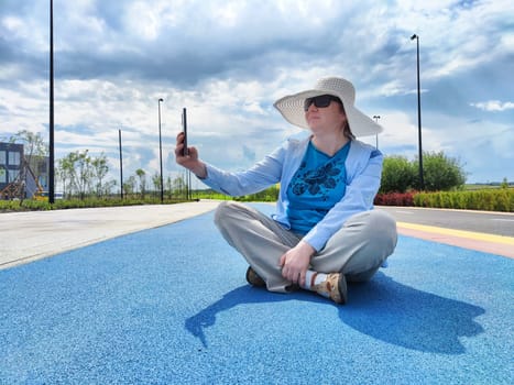 A woman with vitiligo is sitting cross-legged on a colorful pathway, wearing a large-brimmed sun hat and sunglasses while taking a selfie. The sky is partly cloudy.