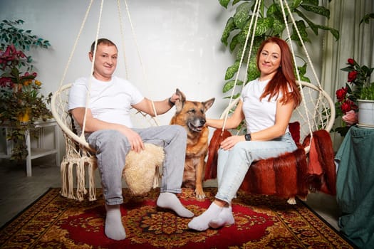 A couple relaxes in their home sitting in hanging chairs, enjoying the company of their joyful German Shepherd dog.