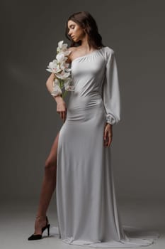 Elegant woman in a white one-shoulder dress holding white orchids, exuding grace, femininity, and sophistication in a minimalist studio setting.