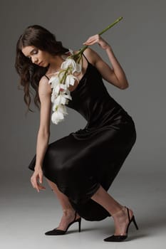 Elegant woman in a black satin dress bending gracefully while holding white orchids, showcasing beauty, fashion, and sophistication in a minimalist studio