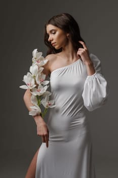 Elegant woman in a white one-shoulder dress holding white orchids, exuding grace, femininity, and sophistication in a minimalist studio setting.