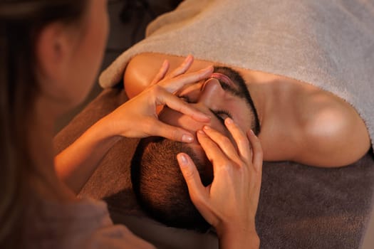 Relaxing facial massage for a bearded man in a spa, performed by a professional therapist. Warm lighting creates a soothing, wellness-focused atmosphere
