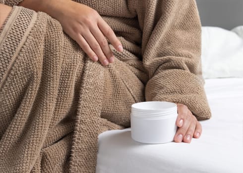 Woman in brown bathrobe puts white opened cream jar on white bedsheet closeup. Bodycare concept. Organic cosmetics for body and face care, beauty and spa procedures
