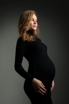 Side view of tranquil pregnant female wearing long black dress and touching tummy while standing on dark background in studio