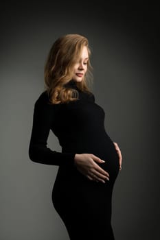 Side view of tranquil pregnant female wearing long black dress and touching tummy while standing on dark background in studio