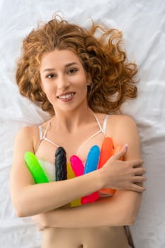 Top view of cheerful female relaxing on soft bed with colorful rubber dildos and looking at camera