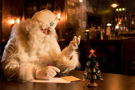 Serious Santa Claus in white hat and costume sitting at table and writing list of obedient kids deserving presents for Christmas