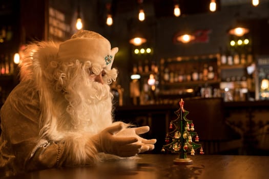Serious Santa Claus in white hat and costume sitting at table and talk with christmas tree