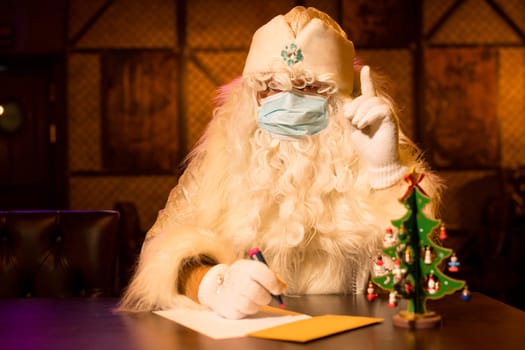 Serious Santa Claus in white hat and protective mask sitting at table and writing a note for Christmas