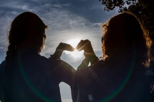 Two individuals form a heart shape using their hands against a bright and beautiful backdrop filled with light