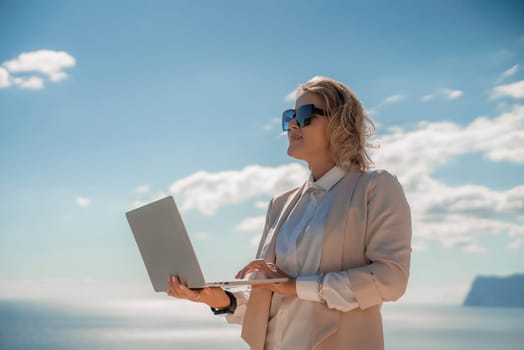 Freelance women sea. She is working on the computer. Good looking middle aged woman typing on a laptop keyboard outdoors with a beautiful sea view. The concept of remote work