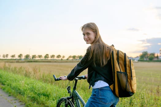 Teenage female with backpack on bike looking at camera, outdoor nature road sunset sky background, copyspace. Activity, lifestyle, youth concept