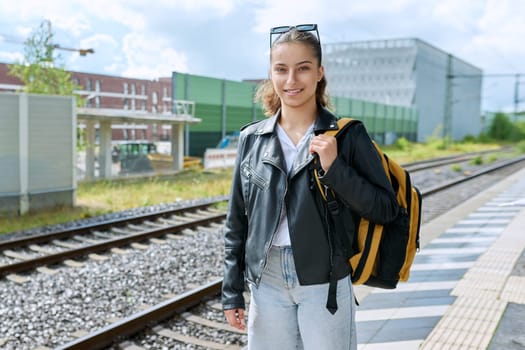Young female waiting for electric train at city railway platform station. Smiling teen girl high school student with backpack. Urban transport, commuter train, passenger transportation, youth