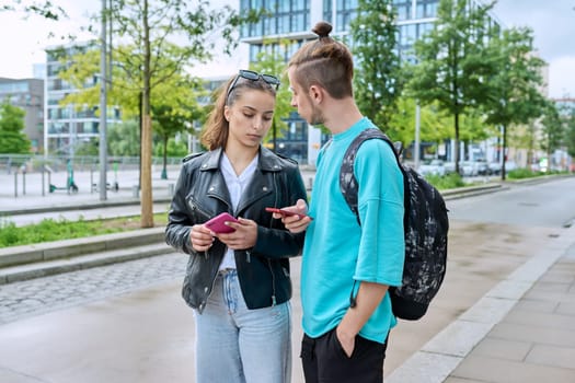 Teen friends guy and girl standing together holding smartphones looking at screen using mobile phones outdoor on city street. Internet digital technology applications for leisure study communication