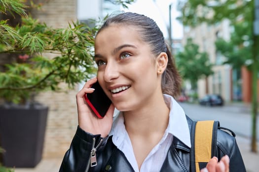 Young attractive teenage girl talking on mobile phone, outdoor on street of modern city. Teenager, college student with backpack, emotionally communicating on smartphone. Urban style, lifestyle, youth