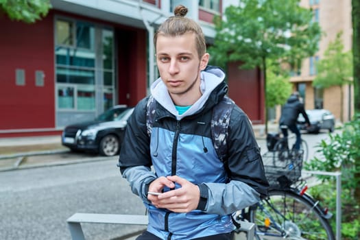 Fashionable young male with backpack using smartphone outdoors in city. Hipster with trendy hairstyle beard with phone in hands mobile applications for tourism travel communication entertainment study