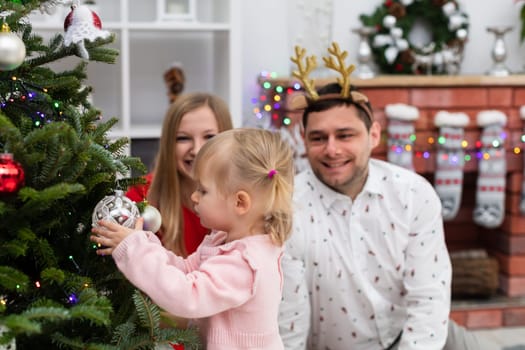 In the foreground a little girl is looking at Christmas tree baubles. A child holds Christmas tree ornaments in her hands. In the background sit smiling parents. The man is wearing a shirt and a headband with reindeer horns on his head.