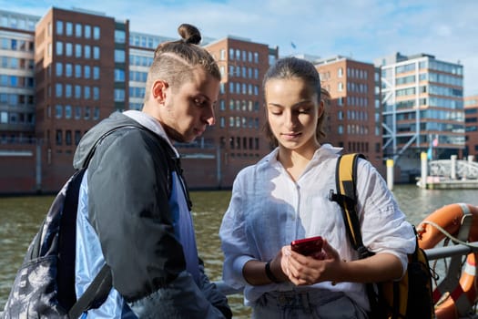 Teen friends guy and girl standing together holding smartphones looking at screen using mobile phones outdoor on city. Internet digital technology applications for leisure study communication