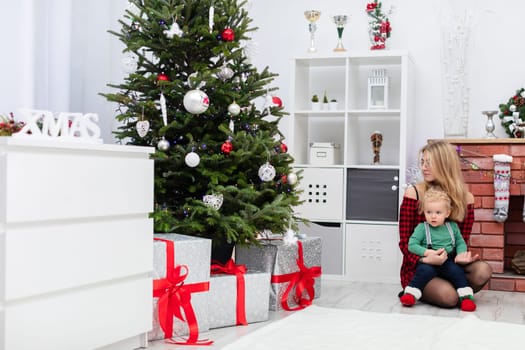 A young blonde woman wearing glasses holds a little boy in a green sweatshirt on her lap. Both are sitting on the floor by the Christmas tree. The Christmas tree is decorated with colorful ornaments. Under the Christmas tree lie large boxes of gifts.