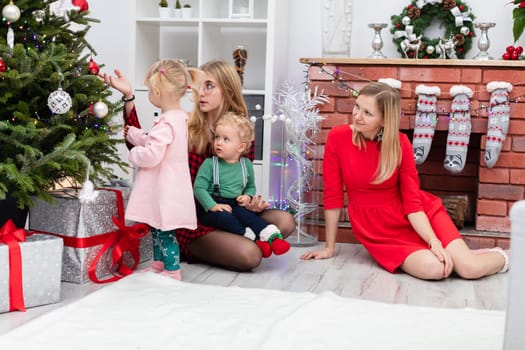 Two adult women and two young children are sitting by the fireplace. One of the women shows the children the baubles hung on the Christmas tree. The house is richly decorated with Christmas decorations.
