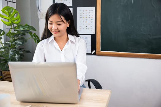 Back to school. Smiling female using computer sitting at school table, Portrait of young woman teacher with laptop at desk in classroom, Online education and learning concept