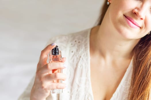 Beautiful young woman with bottle of perfume spraying it on her neck at home.