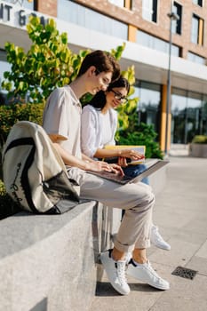 Side view of smiling young students male looking at screen of laptop and female looking down during reading book while sitting together on bench with backpack against blurred green park building