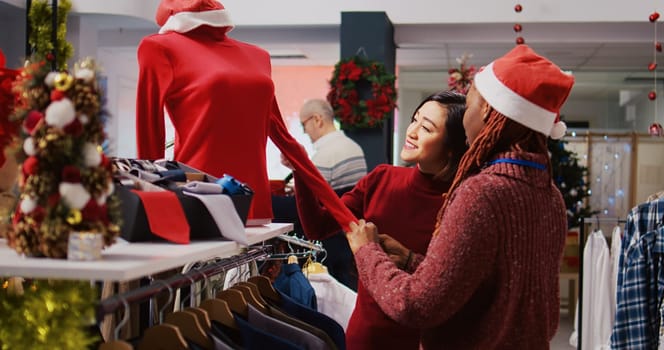 Friendly retail assistant in festive ornate shopping mall fashion shop showing client beautiful red garments, ready to be worn at Xmas themed holiday events during winter season