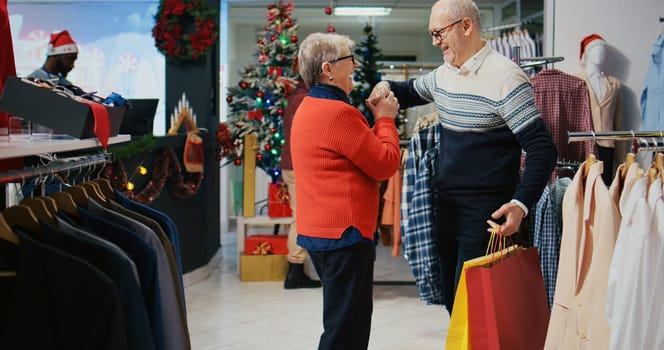 Senior man holding shopping bags, spinning wife in festive decorated fashion shop, happy after finding ideal presents to share with family members at Christmas event party