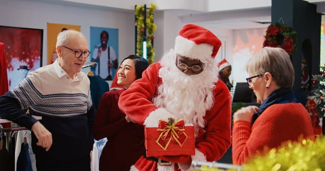 Worker wearing Santa Claus suit holding present box acting as raffle tickets container, mixing them inside in order to pick random winner among excited clients in Christmas ornate fashion shop