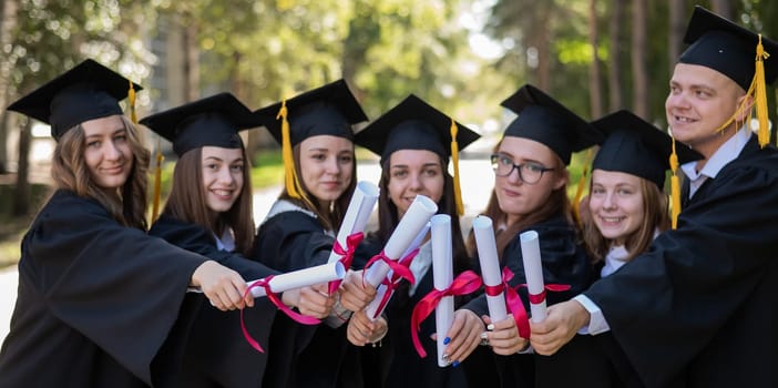 Row of happy young people in graduation gowns holding diplomas outdoors