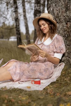 A young beautiful woman in a dress and a round hat reads a book outdoors in the forest and drinks tea. Romantic and vintage photo of a beautiful girl. Reading and relaxation