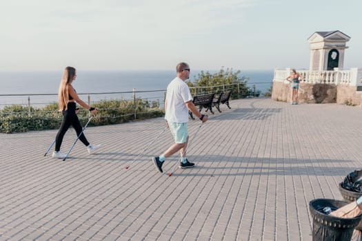 Happy Middle aged couple or friends practicing nordic walking in park near sea. Mature couple with trekking poles walking, practicing Nordic walking outdoors. Aging youthfully and sport concept.